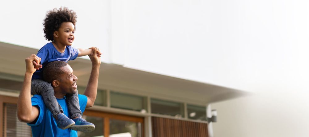 a man holds his child on his shoulders, arms upraised, standing in front of a house that is slightly out of focus and misty, implying having a home is a dream come true. 