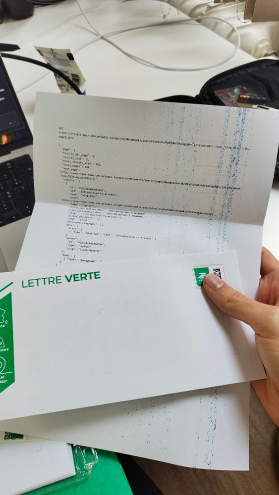Person holding a printed letter with technical JSON API code on it, next to a white envelope labeled “Lettre Verte” from La Poste. The paper has blue ink streaks on the side as if from a printer issue. A laptop and some office items are visible in the background.
