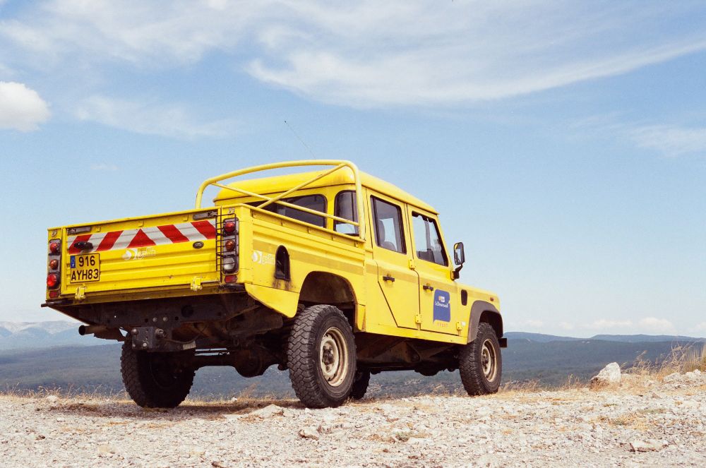 Bright yellow Land Rover Defender pickup parked on a rocky hilltop with a scenic mountain landscape in the background. The vehicle has French license plates and utility markings, suggesting it’s used for off-road or service work. Blue sky with wispy clouds overhead.