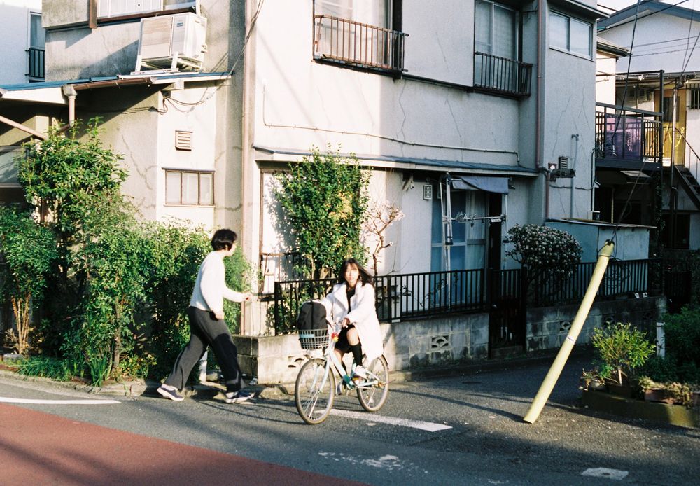 Photograph of two persons, one walking on the left, the other riding a bicycle on the right, both wearing white shirts. The bicycle person appears to be looking over to the right side of the photo for incoming traffic before crossing the street. Behind them, a white-colored house with green bushes is bathed in soft glow from the sun setting.