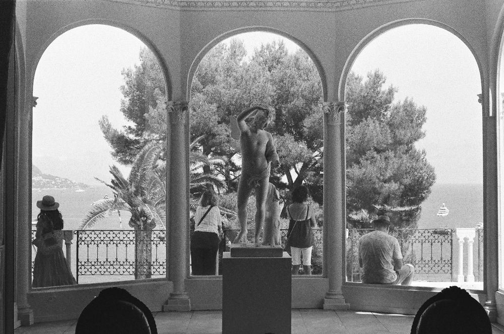 Black and white photo of a classical nude statue framed by three arches, with people standing and looking out at trees and the sea beyond.