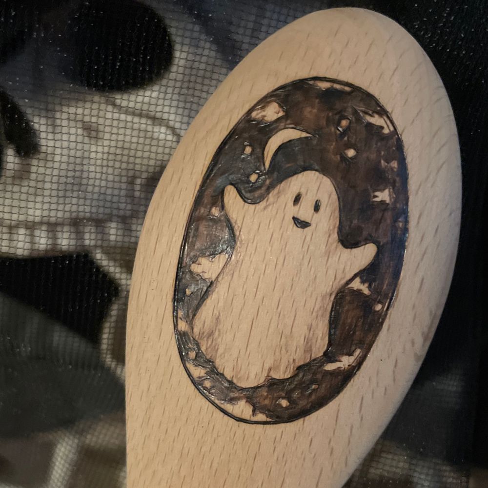 A ghost is wood burned onto a spoon. There’s a skeleton in the background. 