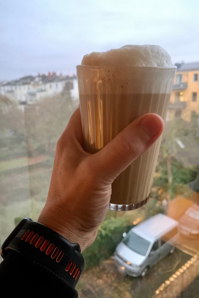Ein Glas Kaffee, gehalten von einer linken Hand vor einem Küchenfenster. Im Hintergrund ein paar rückseitige Häuserzeilen mit ihren Gärten. 