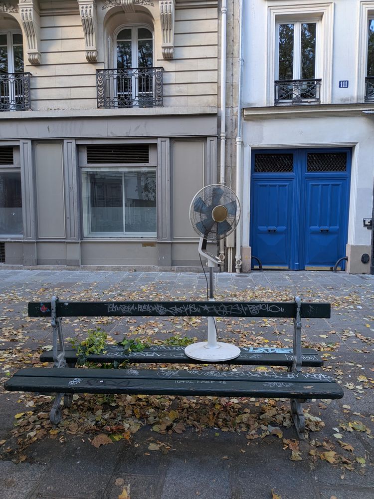 A standing fan stands on a typical parisian bench on a small plaza