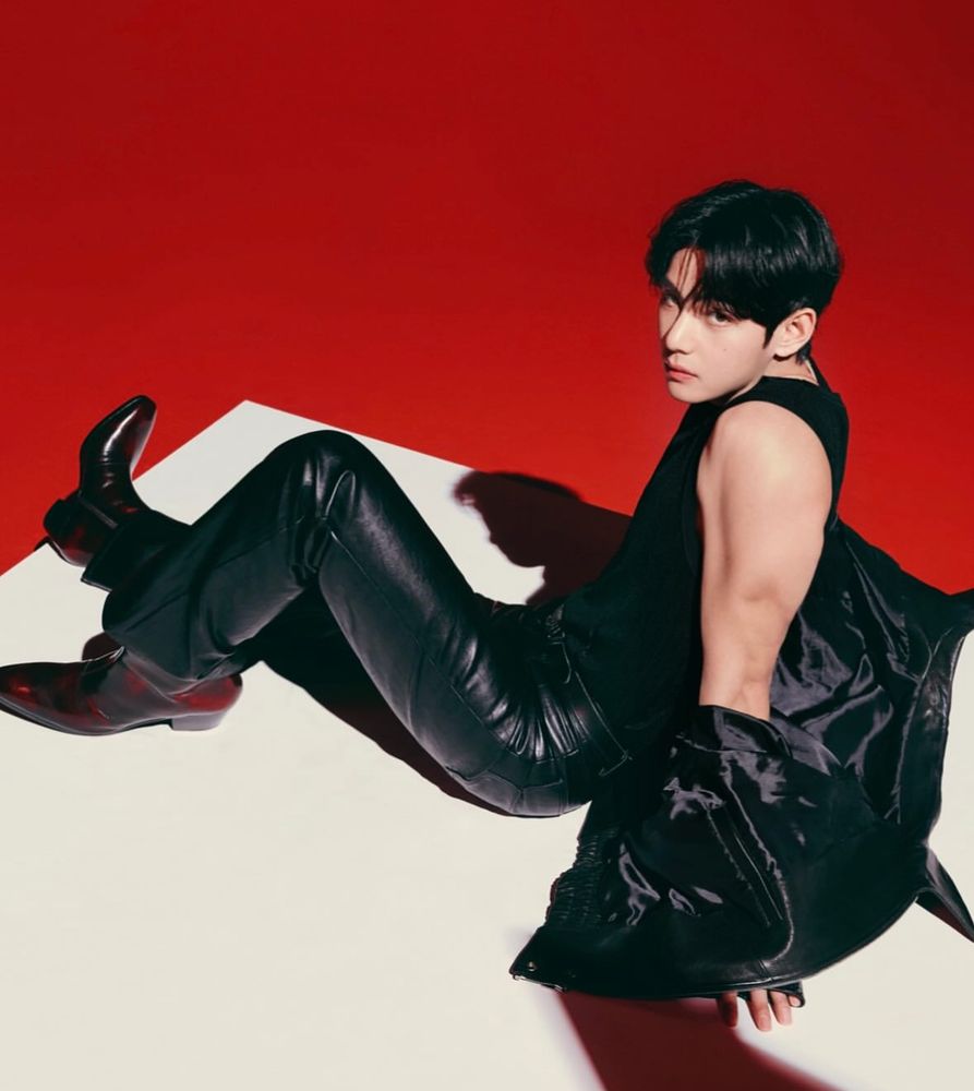 another photo for his tirtir ad. he is wearing all black leather (pants and jacket, dark reddish boots), sitting on a white platform with the background bright red. his head is turned to the audience. he is too good to be true.