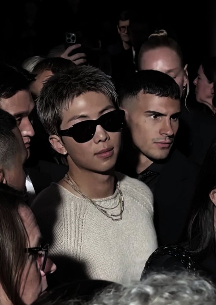 photo of namjoon in the crowd. he is wearing a bottega venetta ensemble (comfy sweater and leather pants), all beige, matching the color of his hair. black sunglasses and some thin chains on his neck. he looks too cool 