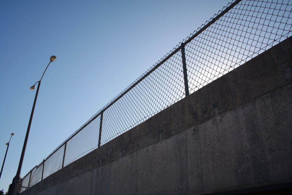 moody chain link fence atop a concrete wall framed on the diagonal and backlight by a cloudless vibrant blue sky, the sun barely masked by the wall 