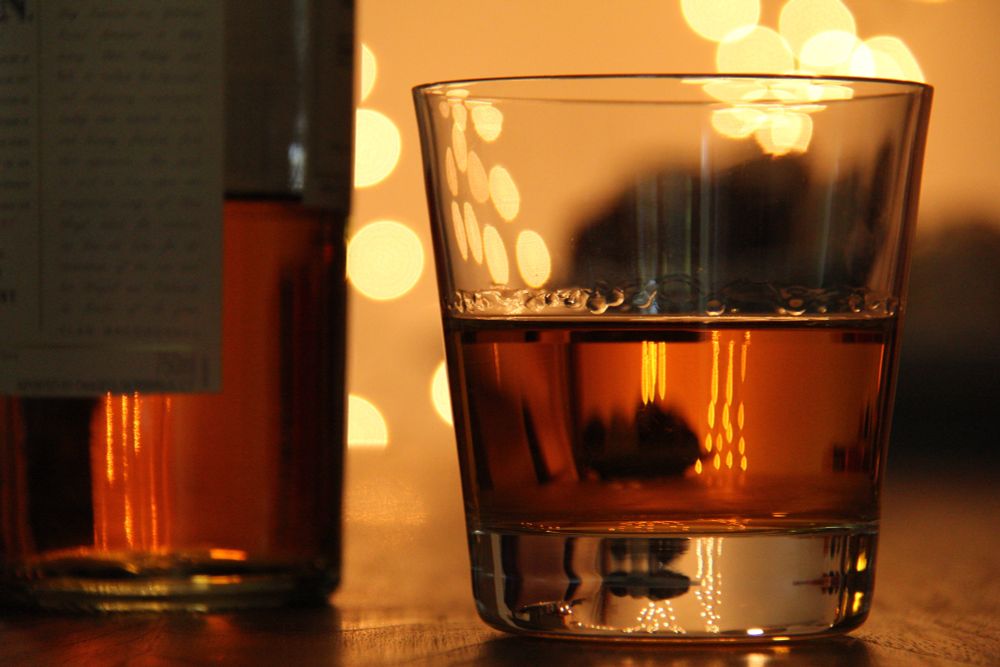 amber and orange, a minimalist tumbler of rich nectar sits on wood alongside a bottle from which it was poured. backlit by glowing yellow lights and peach bokeh