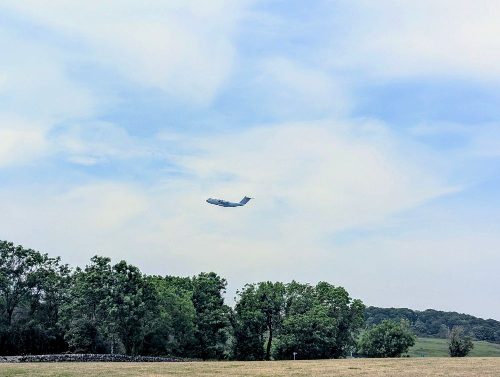 A large RAF plane, Airbus A400M, flying low over the woods outside Newton Stewart today.  Slightly cloudy sky, lots of trees and a bit of the yellow field visible in the photo.