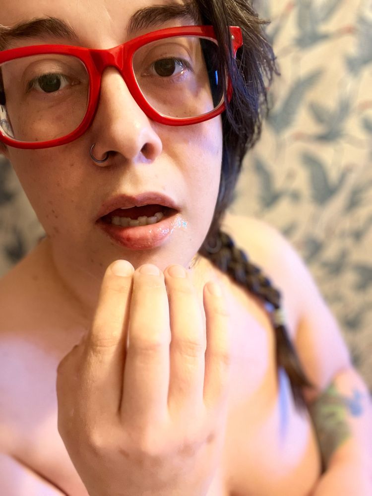 A colorful still photo of a brunette person with brown eyes, a dark nose ring, wearing thick rimmed red and black eyeglasses. They are standing topless in front of a woven shower curtain with blue, red and black printed Sandhill cranes engaging in a courting dance, with their lips pursed open slightly and their right fingers pushed up near their mouth. A sheen of warm saliva and other fluid can be discerned on their bottom lip. Not all fingers taste the same at all times.
