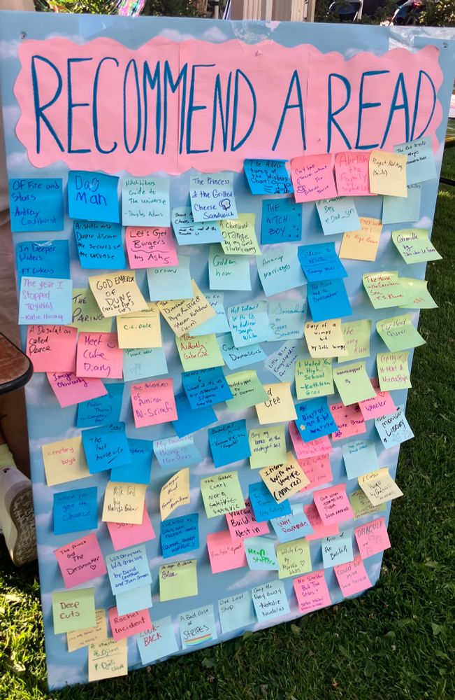 A sign with "recommend a read" on pink paper & about 100 multicoloured post-it notes with reading recommendations stuck to it 