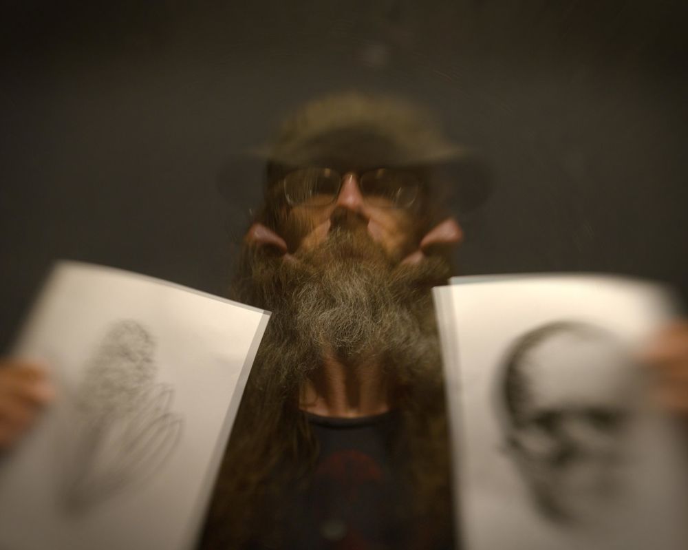A man with a beard stands looking straight ahead. He's holding two sheets of paper, one with a hyacinth on it and one with a skull. Two more versions of him are turned to look at each page. The background is dark grey and indistinct.