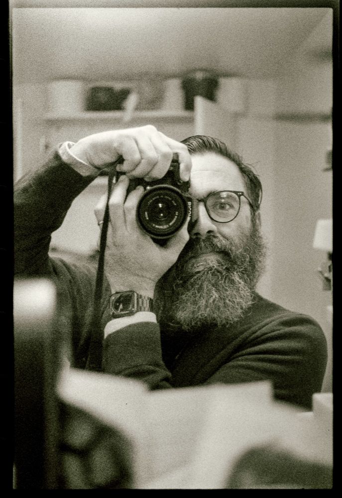 Self portrait in my darkroom mirror. Shelves visible in the background, some stuff on the shelf in the foreground.