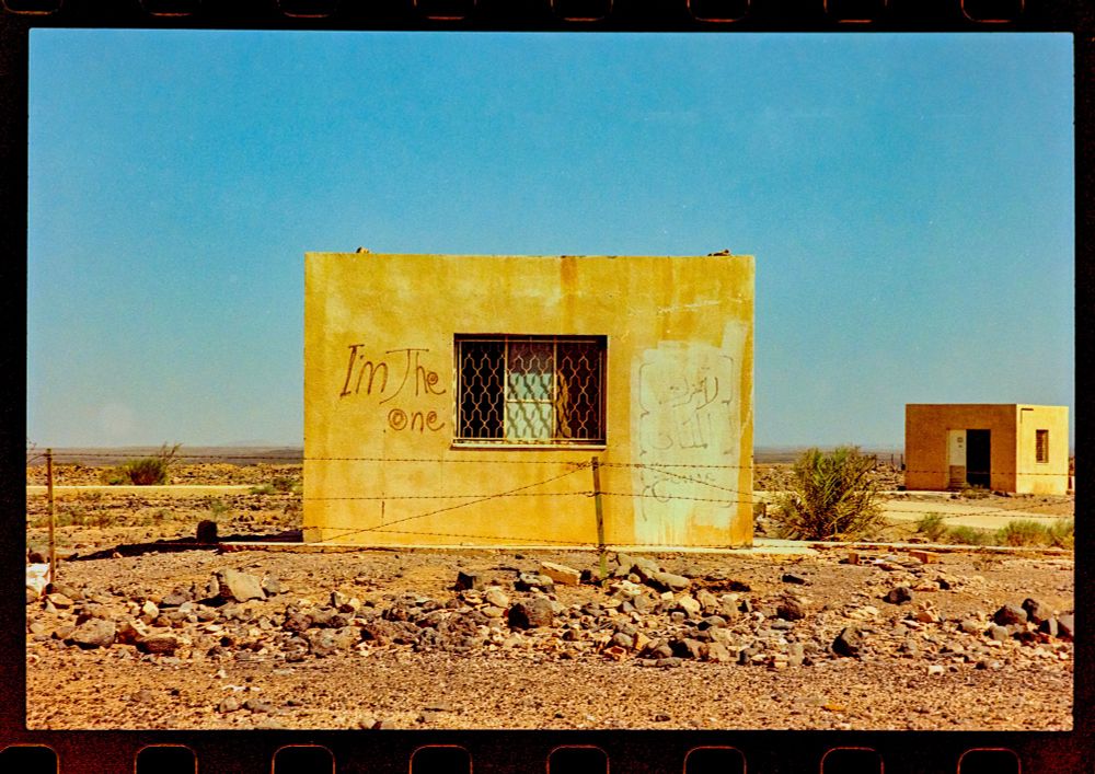 A square building with a fence in front. Graffiti on the building reads “I’m the one”. 