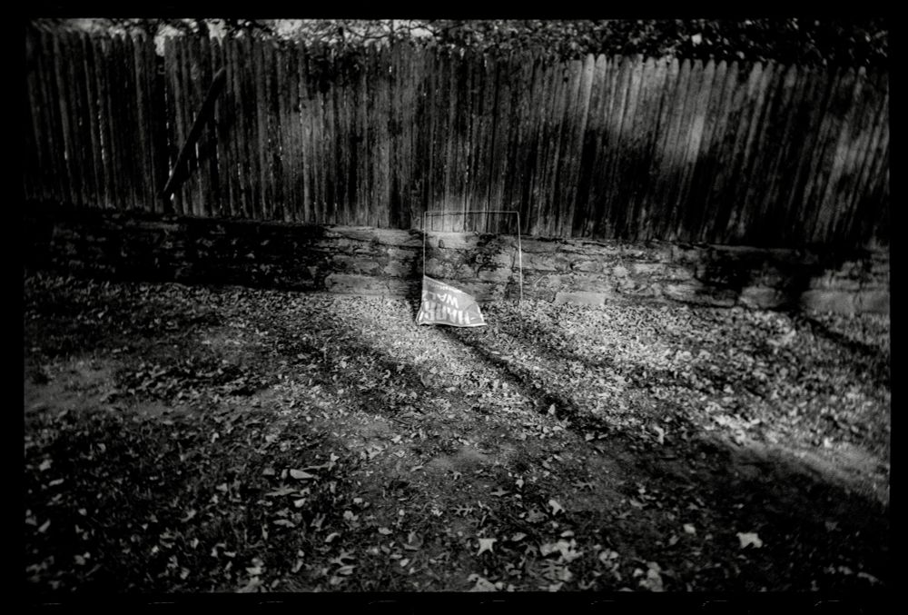 A crumpled and upside down Harris sign in a patch of light leaning against a picket fence among some leaves. 