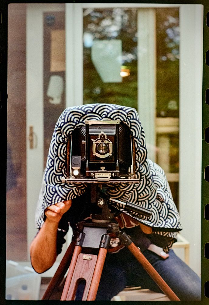 Photo of a man under the darkcloth of a different wooden camera. The camera is pointed directly at the viewer. 