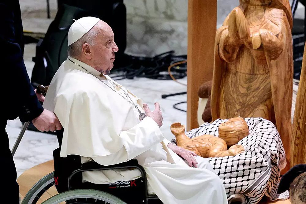 Image of the Pope approaching a nativity scene that features a wood carved baby Jesus who is lying on top of a Palestinian keffiyeh. 
