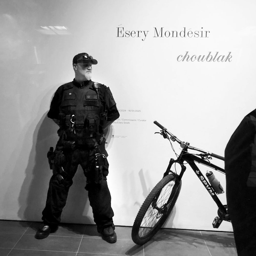 Black and white photo of an armed Montreal police officer standing guard inside the entrance of the Leonard and Bina Ellen Art Gallery - from October 31st, when Concordia University ejected the entire Gallery team from their workplace, in order to use the Gallery to detain students protesting increased police presence on campus. 
