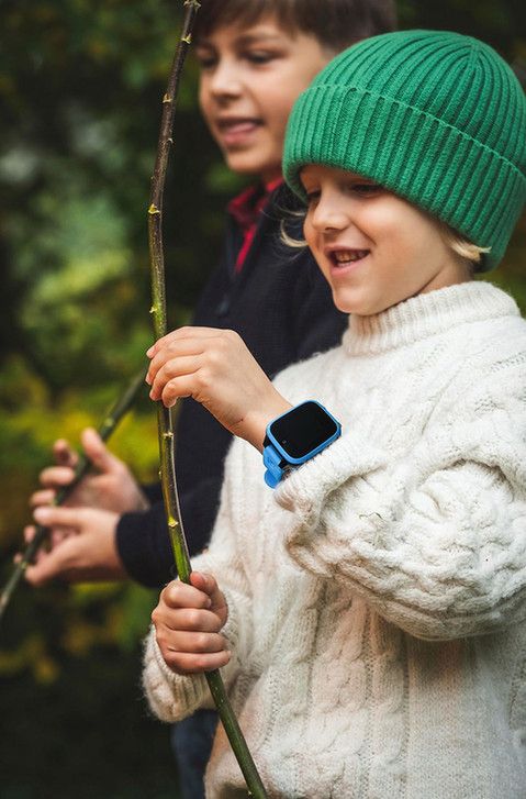 Das Bild zeigt zwei Kinder in einer winterlichen Outdoor-Szene. Im Vordergrund ist ein Kind in einem weißen Strickpullover und einer grünen Strickmütze zu sehen, das eine blaue Smartwatch trägt. Das Kind hält einen langen Ast oder Zweig und lächelt. Im Hintergrund ist ein weiteres Kind zu sehen, das dunkle Kleidung trägt. Die Umgebung ist grün und natürlich, vermutlich in einem Garten oder Park. Die Stimmung des Bildes ist fröhlich und verspielt, und es vermittelt einen Eindruck von Kindern, die die Natur erkunden.