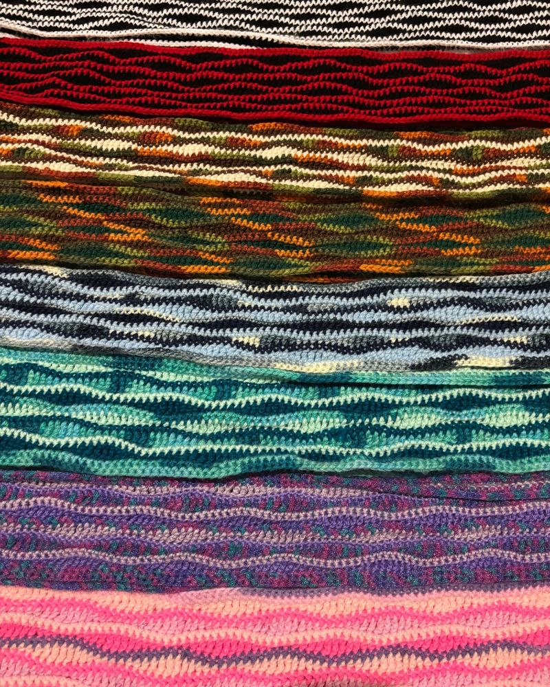 Eight wavy patterned crochet scarves lined up horizontally in a variety of colors, from bright, to autumnal (browns and greens) to black and white. 