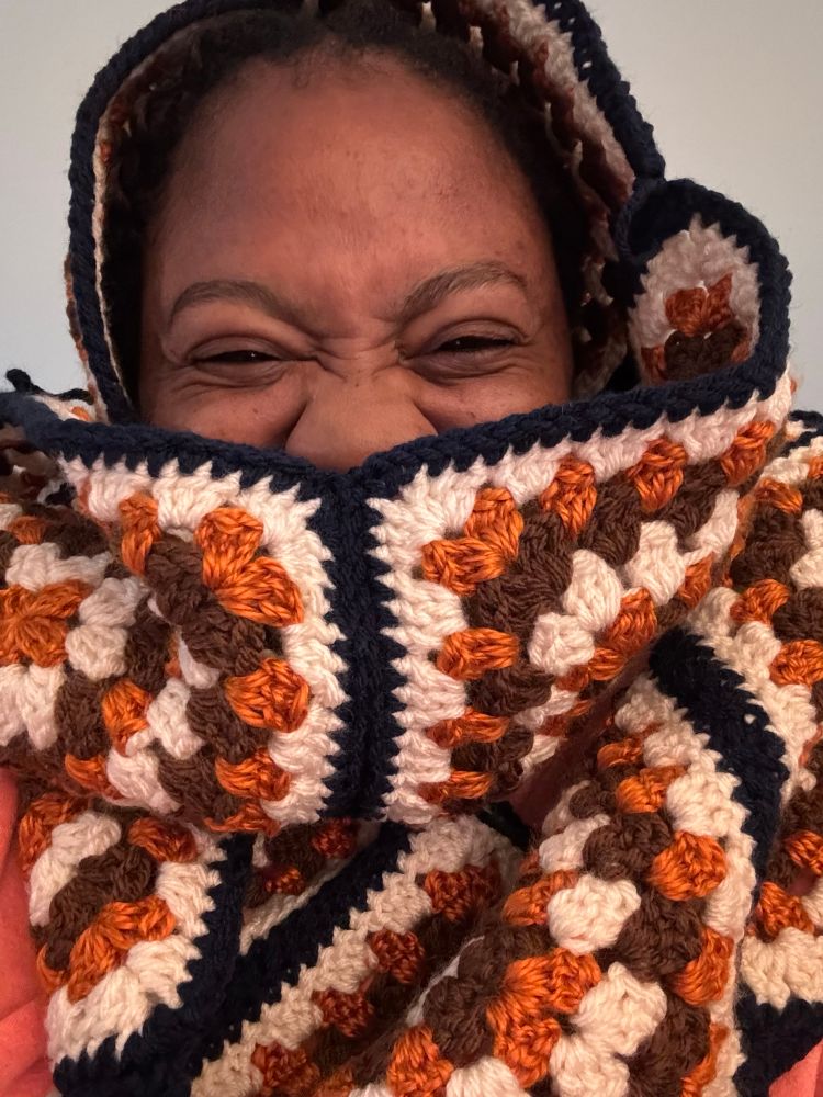 Faye (a young black woman) wrapped in an orange, brown, cream, and navy scarf made of big crochet granny squares. Her face is mostly obscured (only visible from the nose up) her eyes are crinkly as she smiles.