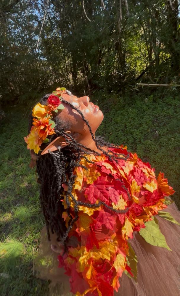 A photo of a young black woman (Faye) outside wearing an autumn fairy costume - her top is made of one long sleeve of maple leaves in shades of red and orange. Her tutu is brown with green leaves along the waistband. She’s wearing dark brown tights and ankle boots. Her hair is in long black twists and a crown of autumn leaves and flowers adorns her head. 

She tilts her head up to the sun, a serene smile on her face!