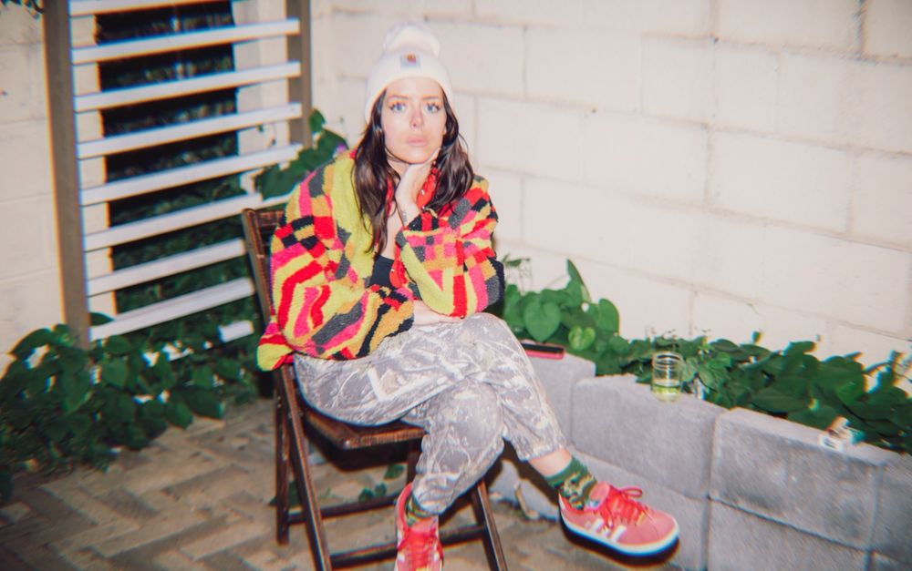 kayla sitting cross-legged in a brown folding chair on a concrete patio at night. same white carhartt hat and multi colored jacket, but now you can see she is also wearing real camo pants, socks that are a different camo and red low top sneakers. her fit is pure chaos. weeds line the periphery of the patio. a glass of wine and a pack of cigarettes sit on a ledge. 