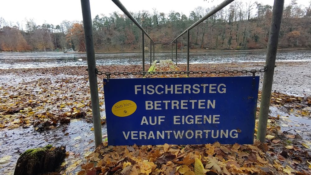 Ein morscher Fischersteg mit viel Laub, daran eine Kette mit einem blauen Schild "Fischersteg Betreten auf eigene Verantwortung". Darauf klebt ein gelber Sticker mit schwarzer Beschriftung, die lautet "Nett hier. Aber waren Sie schon mal in Ankh-Morpork? Stadt der tausend Überraschungen"