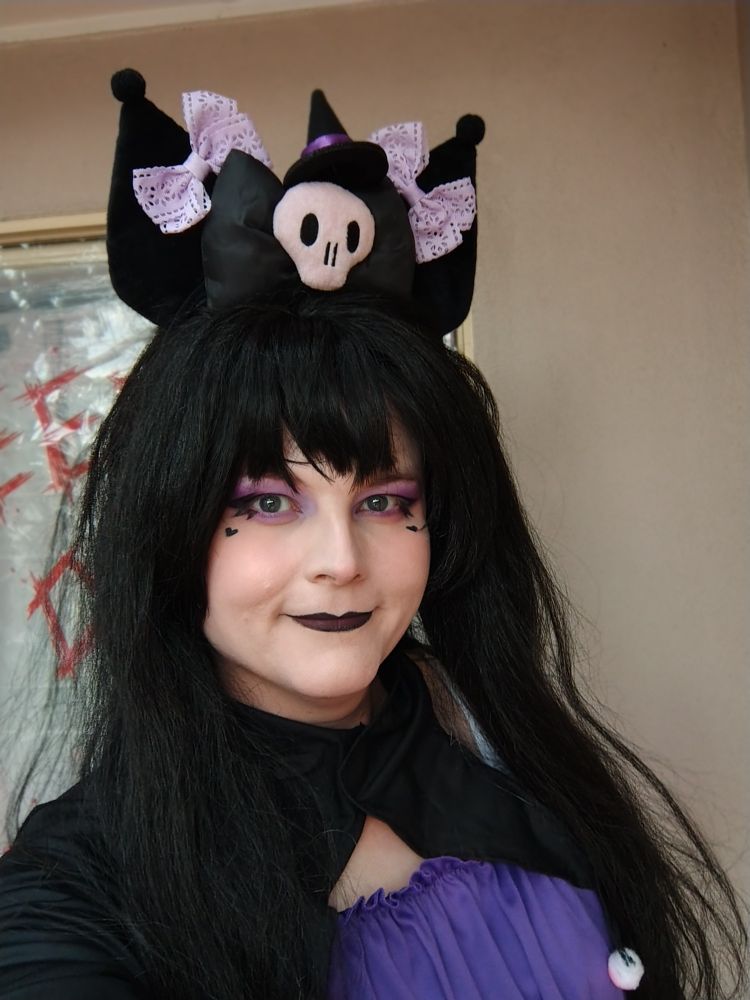 Myself Roxy with cute makeup with a Kuromi headband with purple bows and a little witches hat on top, smiling at camera.