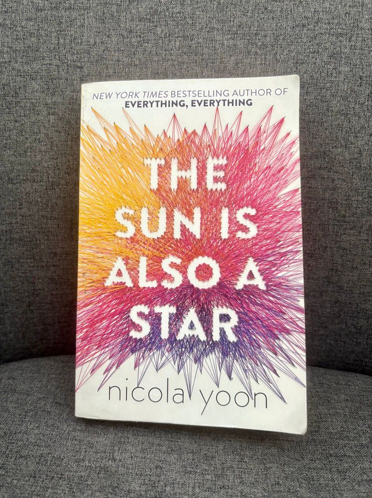 The Sun is also a Star by Nicola Yoon book cover on a grey chair. The title is in white block capitals with yellow pink and purple lines bursting out behind 