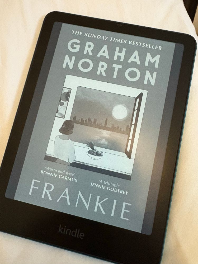 Kindle book cover of Frankie by Graham Norton. Shows a woman looking out of an open window over the sun setting on New York City skyline 