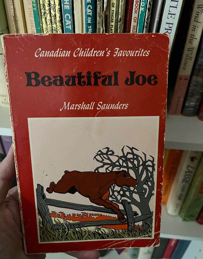 The cover of the children’s novel Beautiful Joe by Marshall Saunders, showing an illustration of a medium-sized brown dog of mixed breed, with a short docked tail and cropped ears, jumping over a wooden fence in a field. A tagline at the top of the cover reads “Canadian Children’s Favourites”.
