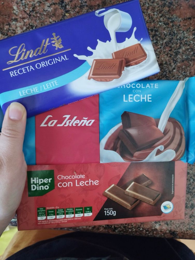 Hand holding 3 large bars of chocolate. 
Lindt receta original leche. La islena chocolate con leche. And hiperdino own brand chocolate con leche
