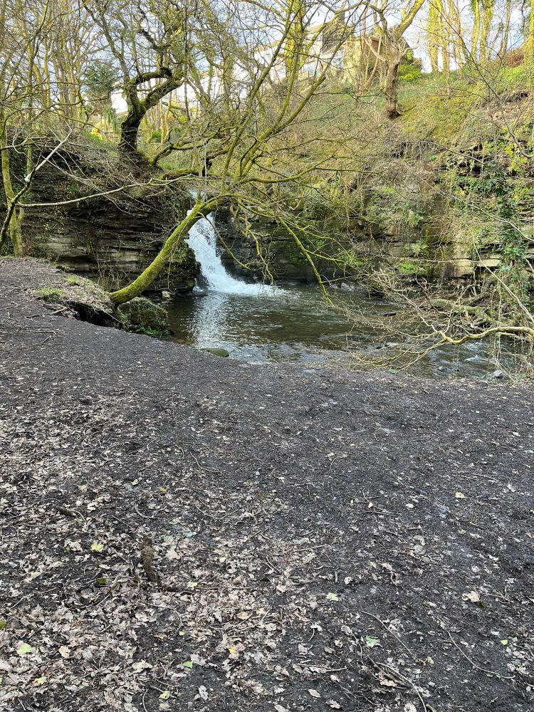A waterfall over rocks in a green and wooded valley.  The ground is gravel