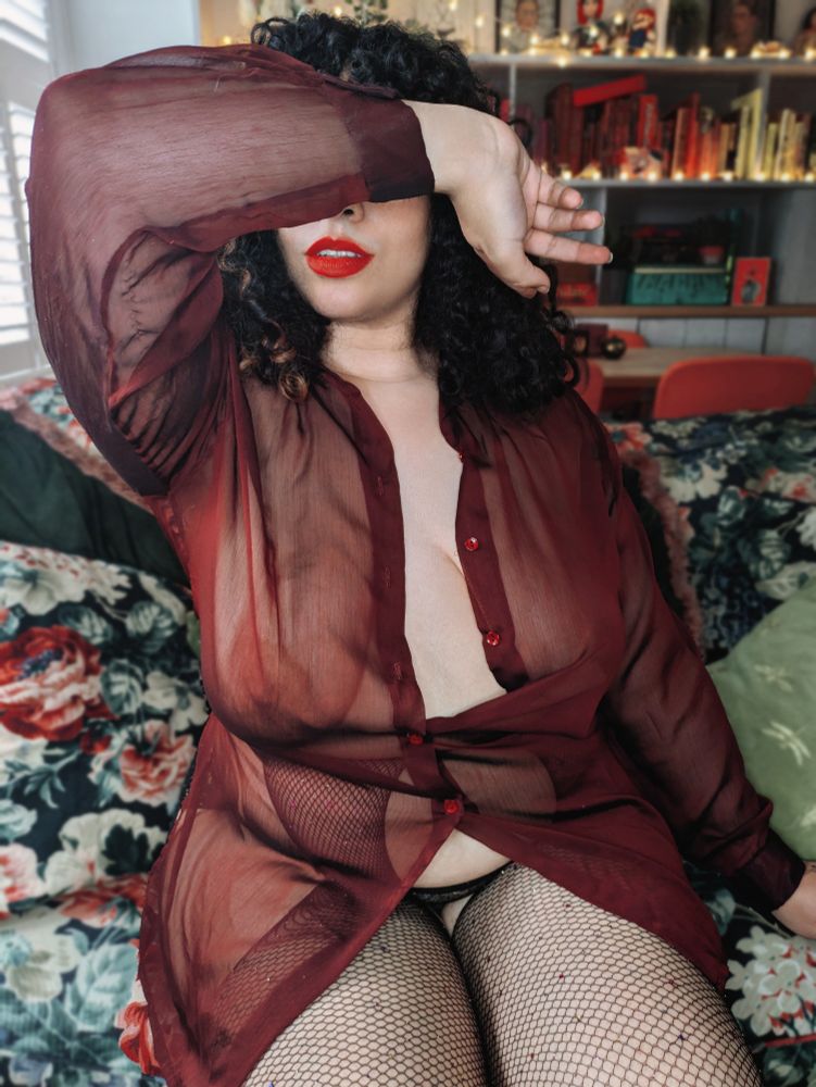 Miss Annie sits on a floral green sofa wearing a  sheer burgundy shirt and fishnets. You can just make out a nipple through the material. She drapes her arm across her face so we only see her red lips. In the background are fairy lights and a bookcase. 