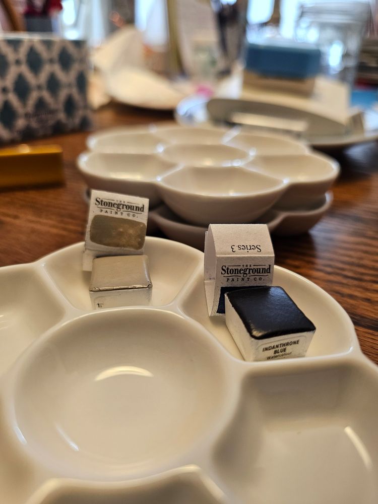 Blurry painting  supplies at top.  Two half paint pans with Stoneground Paint Company wrapping behind them are in wells of ceramic dish. One the left is a putty colour ama on the right is a dark blue colour. 