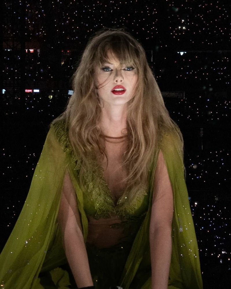 Taylor Swift in her green folklore outfit performing at the Eras Tour looking at the camera with under lighting.