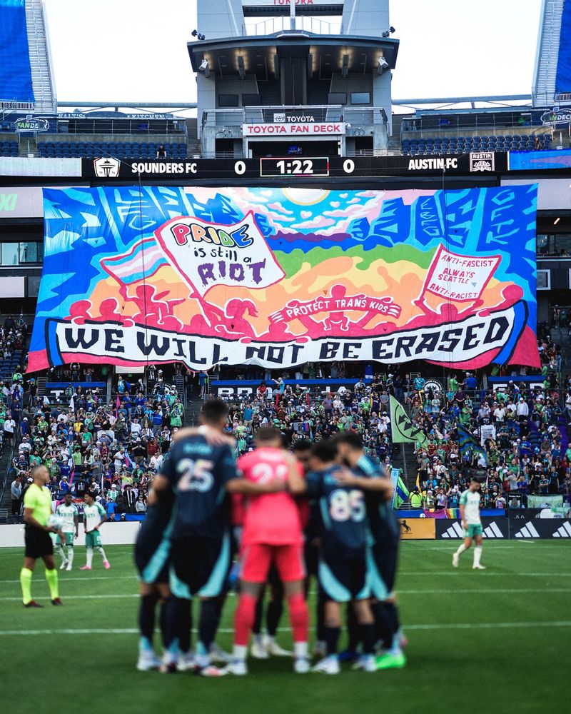 ECS tifo that says "Pride is a riot" and "We will not be erased" -- Other elements include a "protect trans kids" scarf, a trans flag, and a two-pole that says "anti-fascist anti-racist always Seattle"