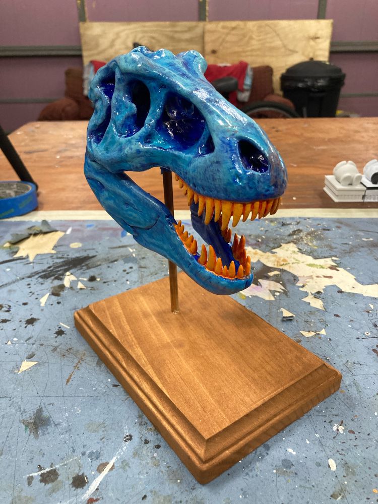 A small blue Tyrannosaurus Rex replica with orange teeth on a wooden stand. The view is 3/4 from the front