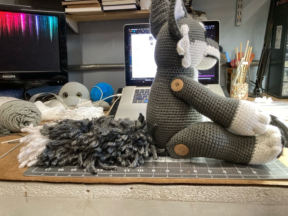 Gray and white crochet wolf sitting at profile view on a desk with a big fluffy tail