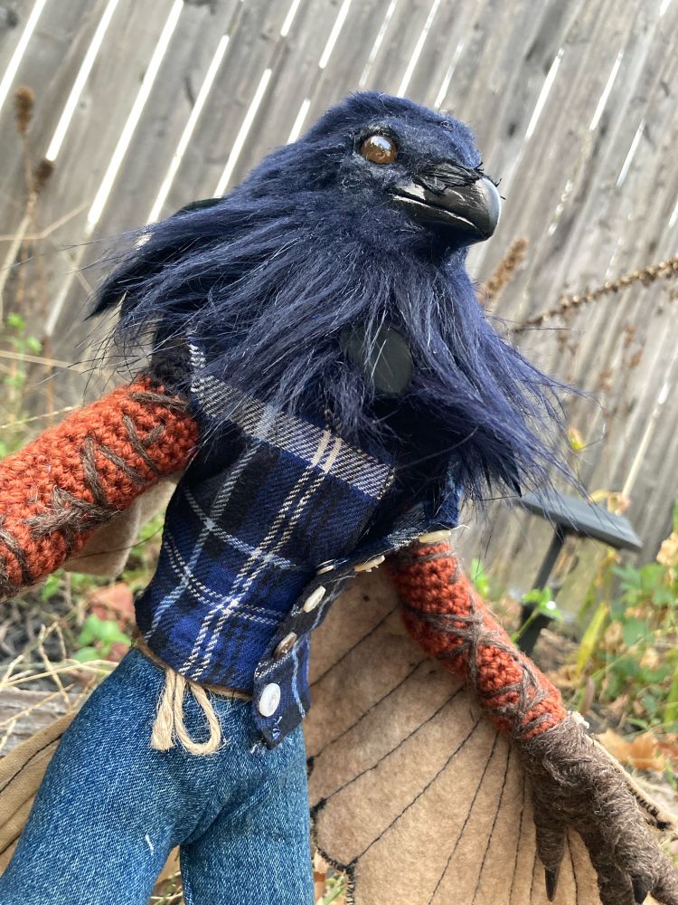 A crow art doll standing outside but a close up from below, the doll is an anthropomorphic crow art doll about 18 inches tall. Its arms and legs are dark are wrapped in brown yarn, the arms have large tea stained off white wings coming off them. The clothes are a blue flannel shirt and jeans made of old clothes. 