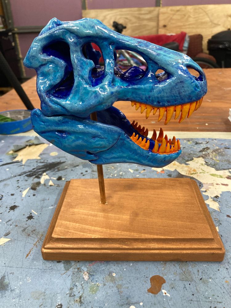 A small blue Tyrannosaurus Rex replica with orange teeth on a wooden stand. The view is of its profile