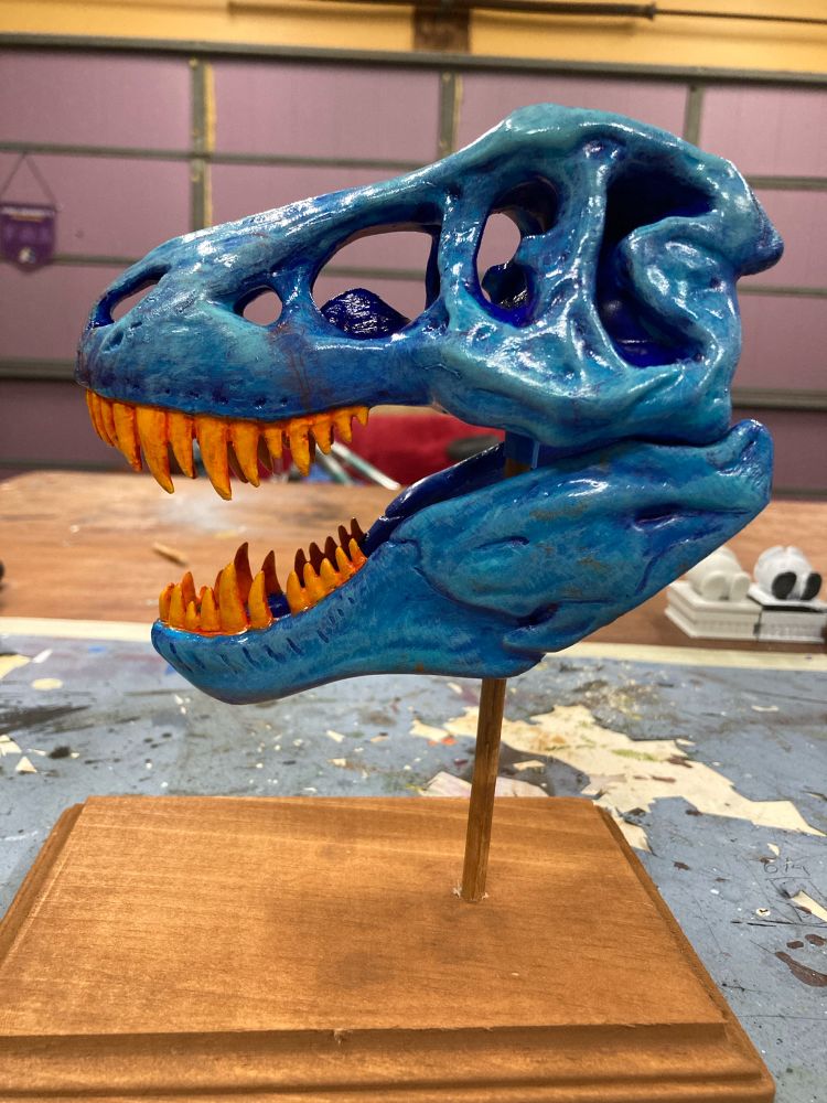 A small blue Tyrannosaurus Rex replica with orange teeth on a wooden stand. The view is of its profile. 