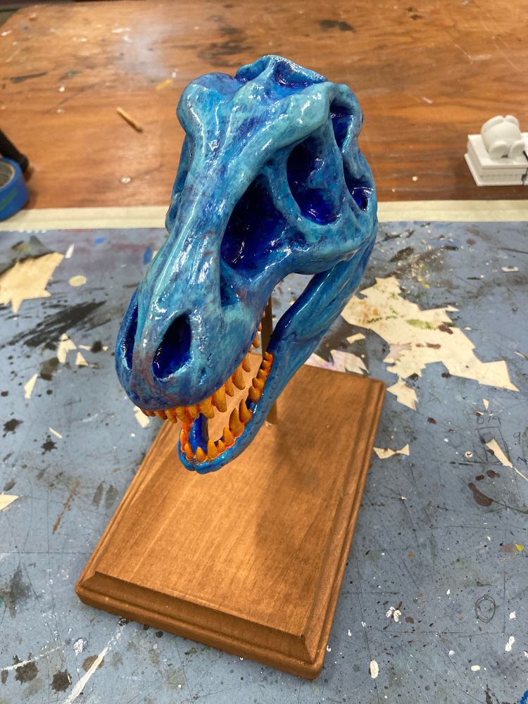 A small blue Tyrannosaurus Rex replica with orange teeth on a wooden stand. The view is from above
