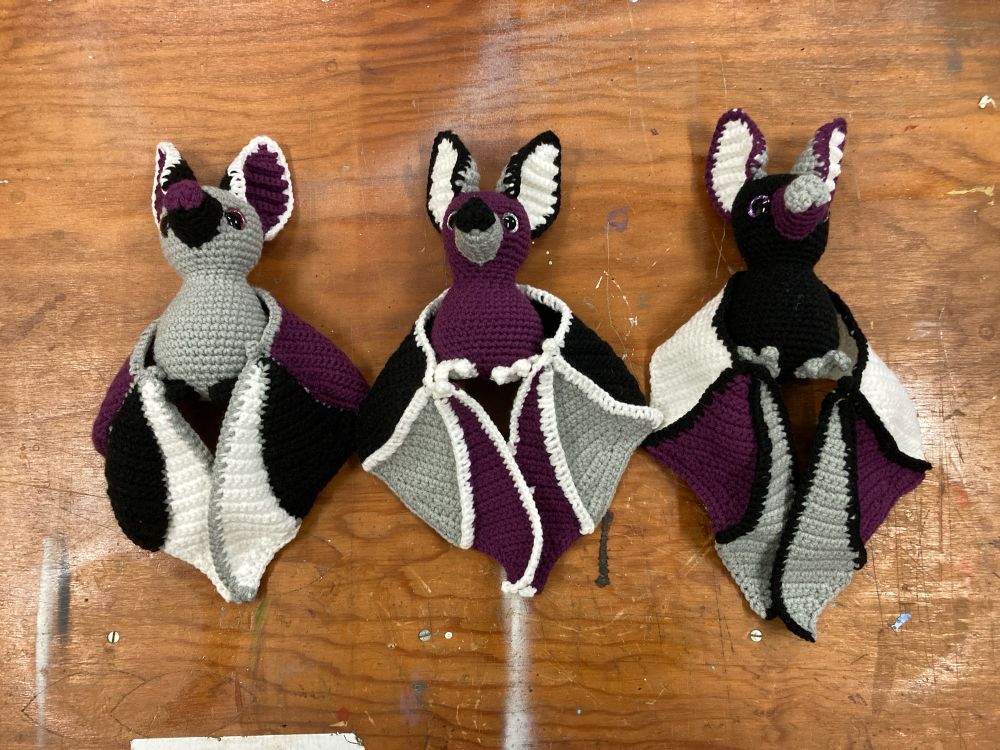 3 crochet bats in Ace pride colors on a wooden table top. Laying down with wings folded.