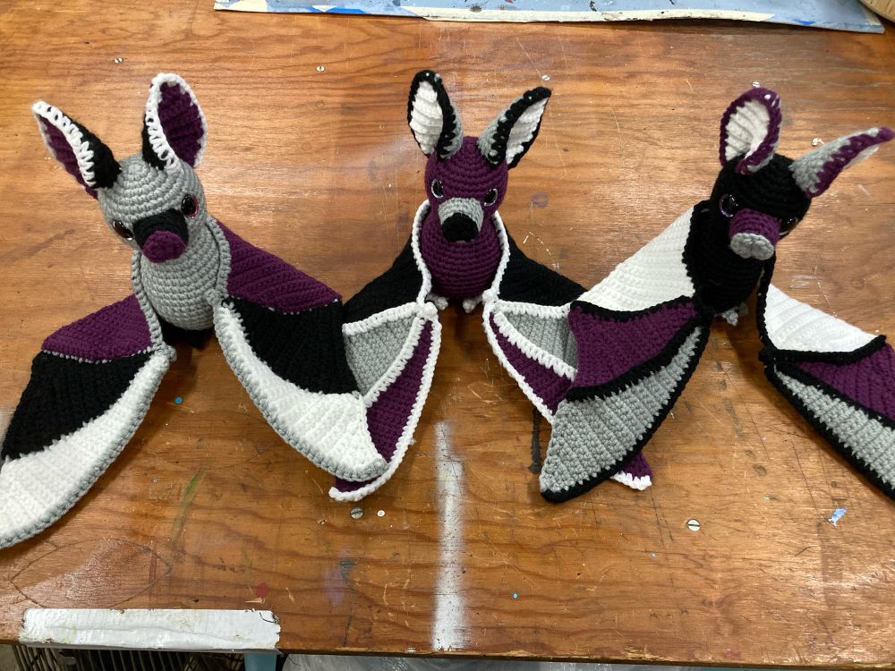 3 crochet bats in Ace pride colors on a wooden table top.