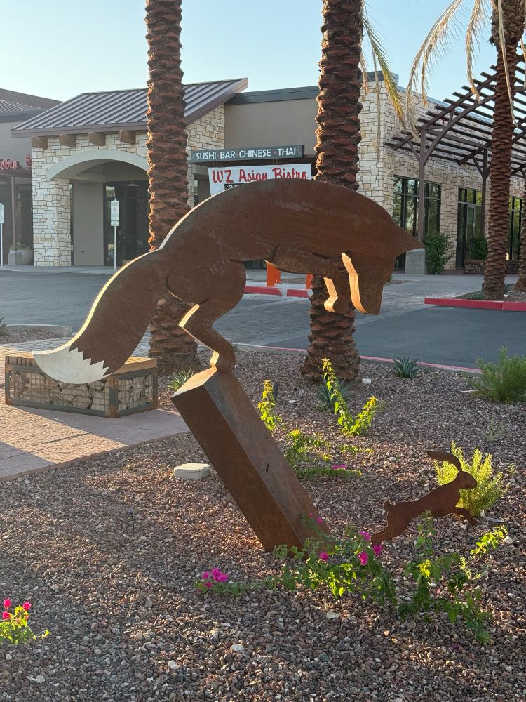 Statue of a coyote perched on a leaning obelisk who is about to pounce on a leaping jackrabbit.