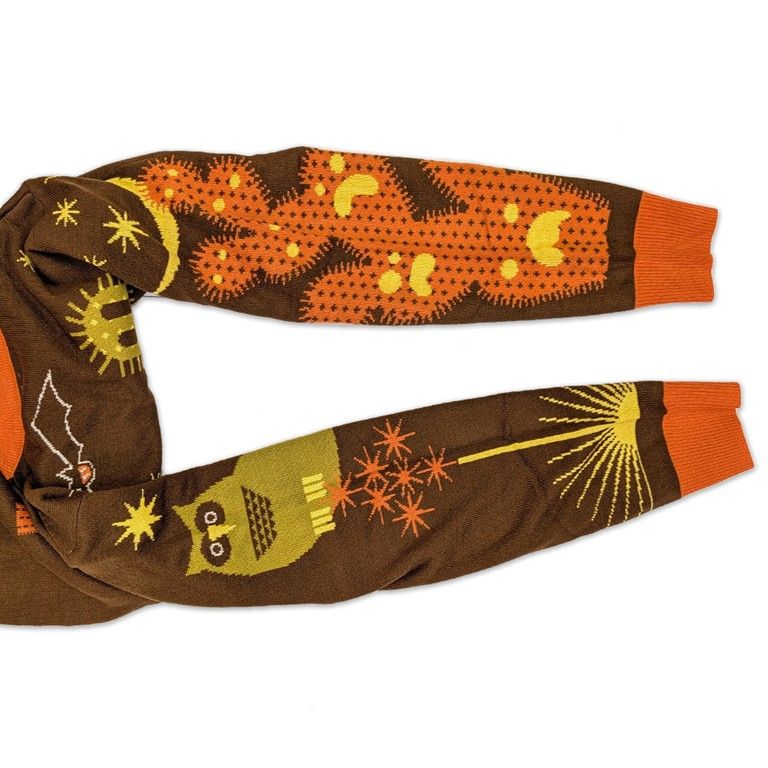 The Night Vale Fall Sweater has an orange cactus with unhappy pumpkin faces cut out going down the right sleeve. On the left sleeve is an owl with a yellow star above, many smaller orange stars below it and a yellow sun with long rays at the bottom, near the orange cuff. 