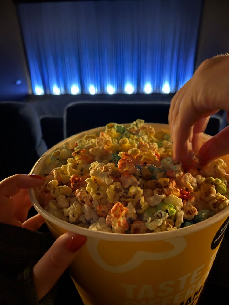 Herr Drops und meine Hände in einem Popcorneimer, im Hintergrund eine noch mit Vorhang verschlossene Kinoleinwand in leerem Kinosaal.