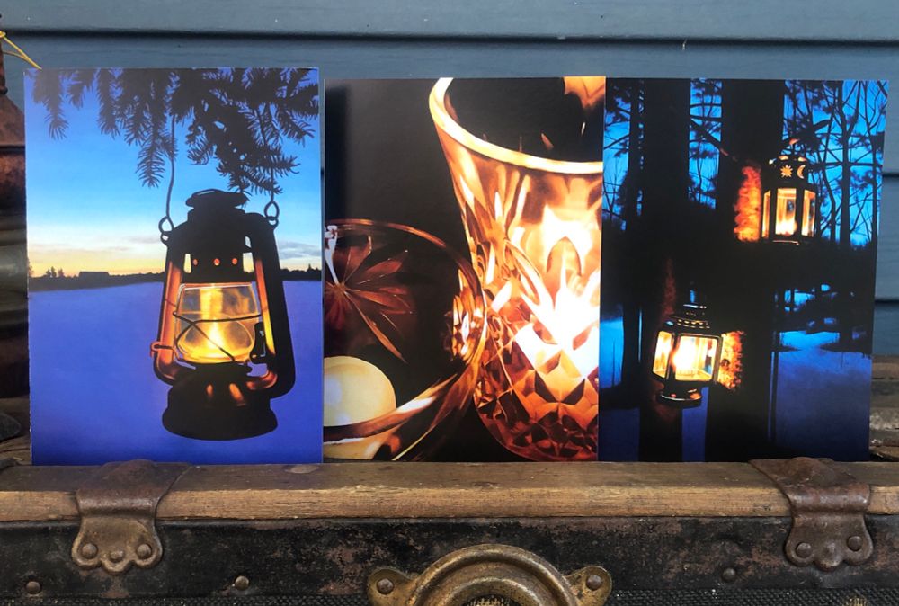 3 art cards standing up on an old shipping trunk. One is an oil lamp hanging from tree branches, one is a close up of candles in crystal holders, and one is 2 lanterns hanging from a tree in dark woodland.